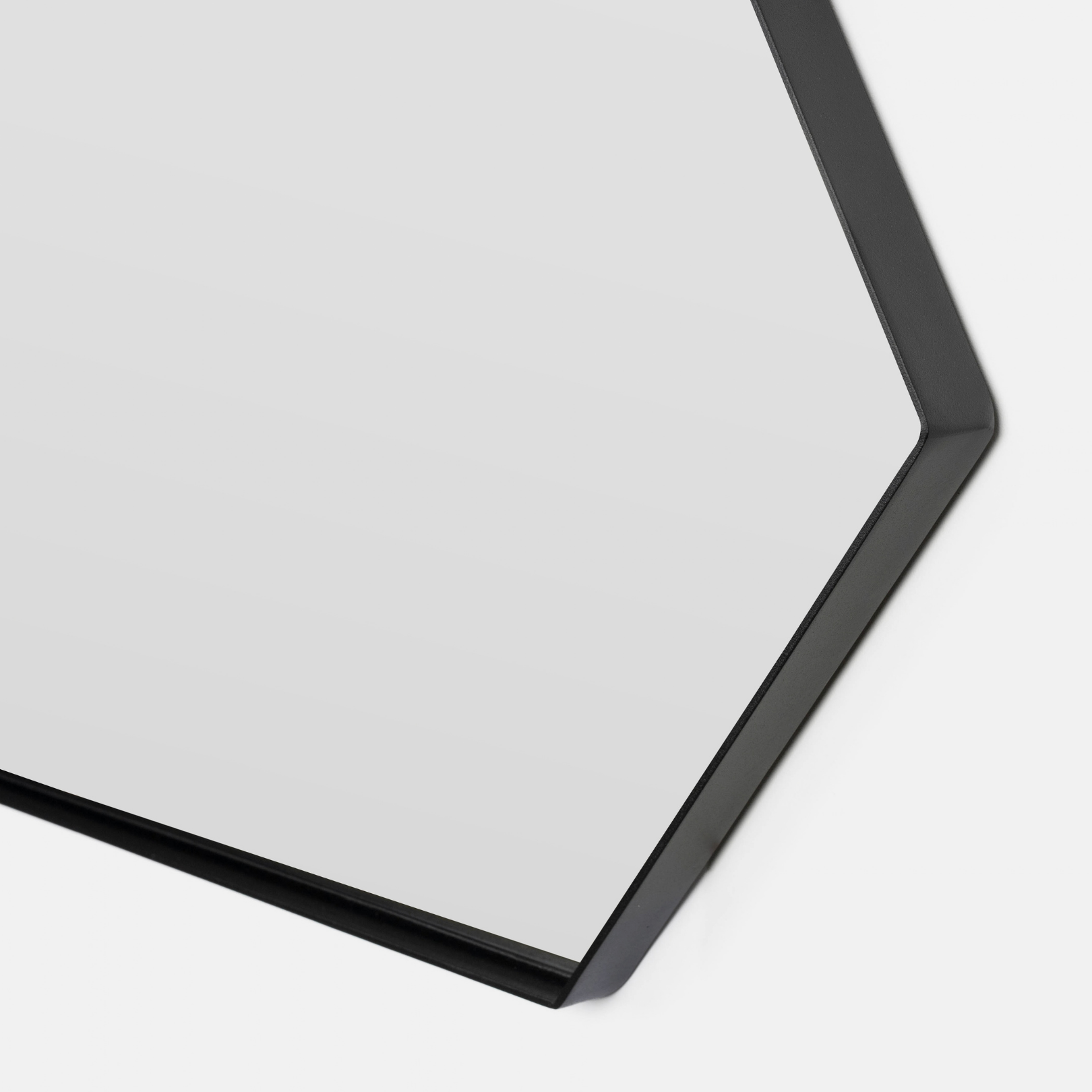 Close-up of a hexagonal wall mirror with a slim black frame, showing a portion of the reflective glass surface and its corner against a light gray background.