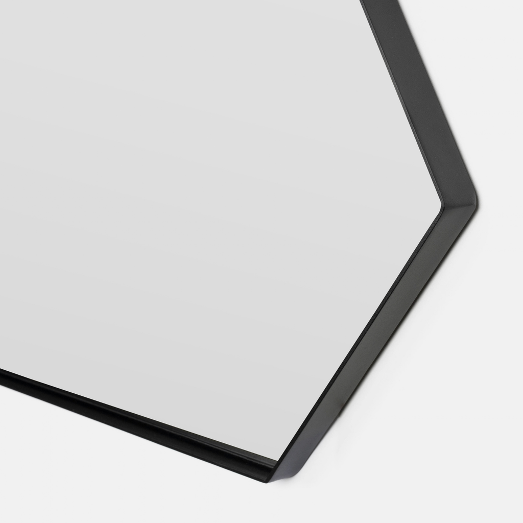 Close-up of a hexagonal wall mirror with a slim black frame, showing a portion of the reflective glass surface and its corner against a light gray background.