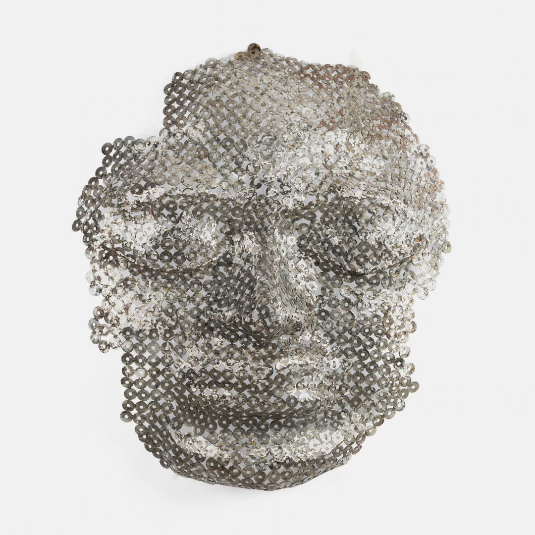 A sculptural metallic mask made of densely packed silver and gray sequins, photographed against a plain light gray background.