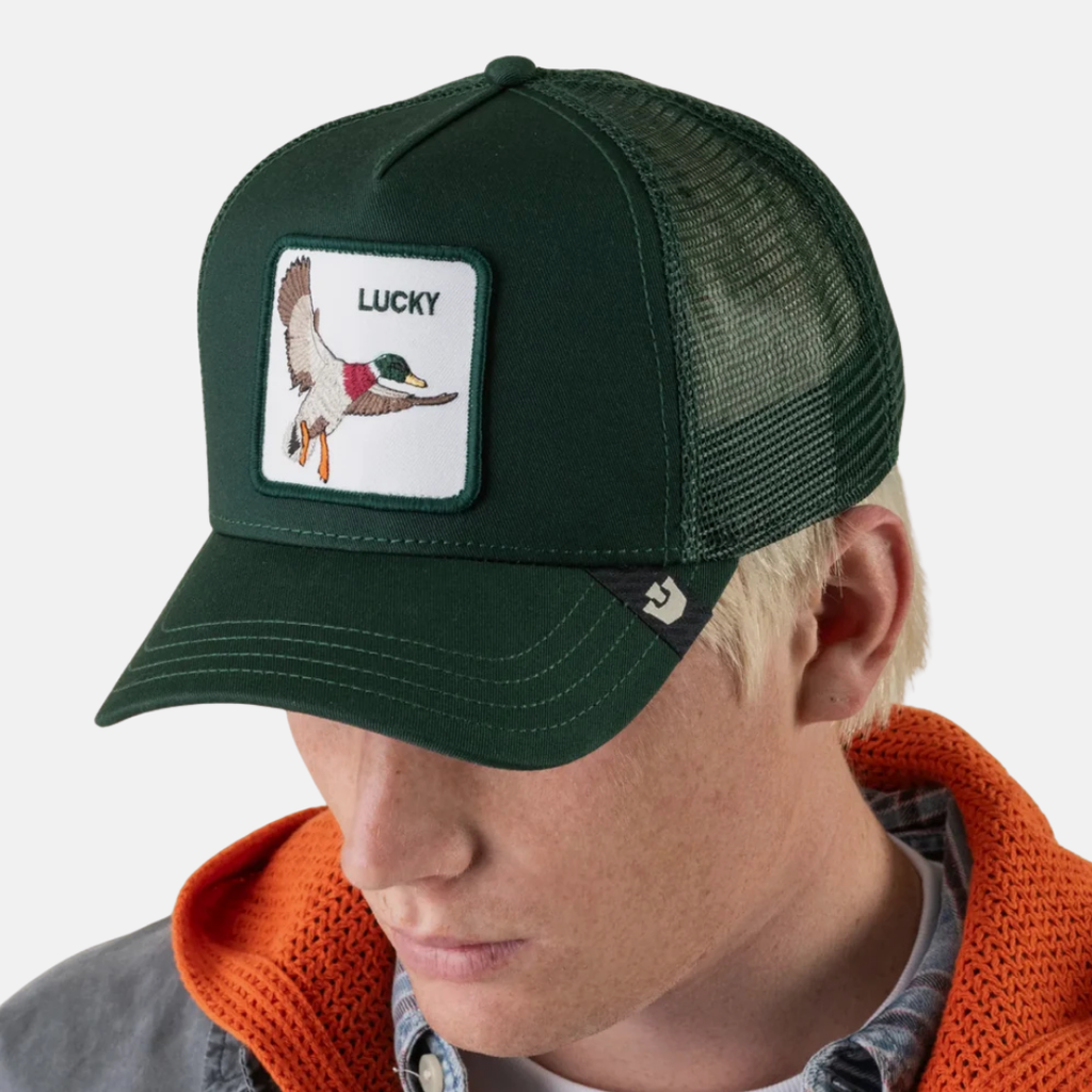 A dark green trucker hat with a white rectangular patch featuring a duck illustration and the word 'LUCKY', worn by a model in an orange hoodie against a light gray background.